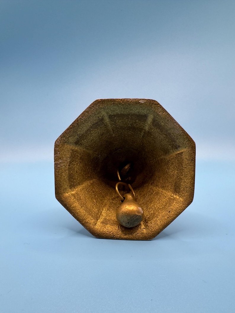 K&ouml;nnte beinhalten: Vintage Messingglocke mit achteckiger Oberseite und kleinem Kl&ouml;ppel. Die Glocke hat ein angelaufenes, gealtertes Aussehen mit einem gr&uuml;nlichen Farbton in einigen Bereichen. Der Hintergrund ist einfarbig hellblau.