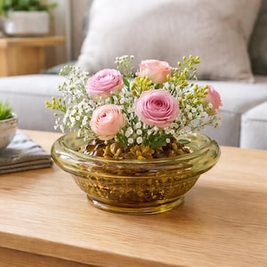 May include: An amber-coloured glass bowl filled with pink roses, white baby's breath, and greenery. The bowl has a textured design and sits on a wooden table. The floral arrangement is a decorative centrepiece.
