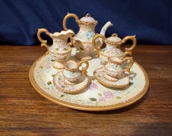 May include: A miniature porcelain tea set with a floral design. The set includes a teapot, creamer, sugar bowl, and two teacups with saucers, all arranged on a matching tray. Gold trim accents the edges.