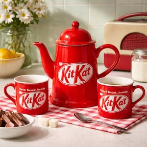 Puede incluir: Una tetera roja de Kit Kat y dos tazas a juego se encuentran sobre un paño a cuadros rojos y blancos. Las tazas y la tetera tienen el logotipo de Kit Kat en blanco. También hay un cuenco con chocolate y terrones de azúcar.