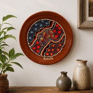 May include: A round, brown decorative plate with an Aboriginal art design featuring a kangaroo in the center. The kangaroo is depicted in red, yellow, and black, surrounded by dots in various colors. The word "Australia" is written at the bottom.