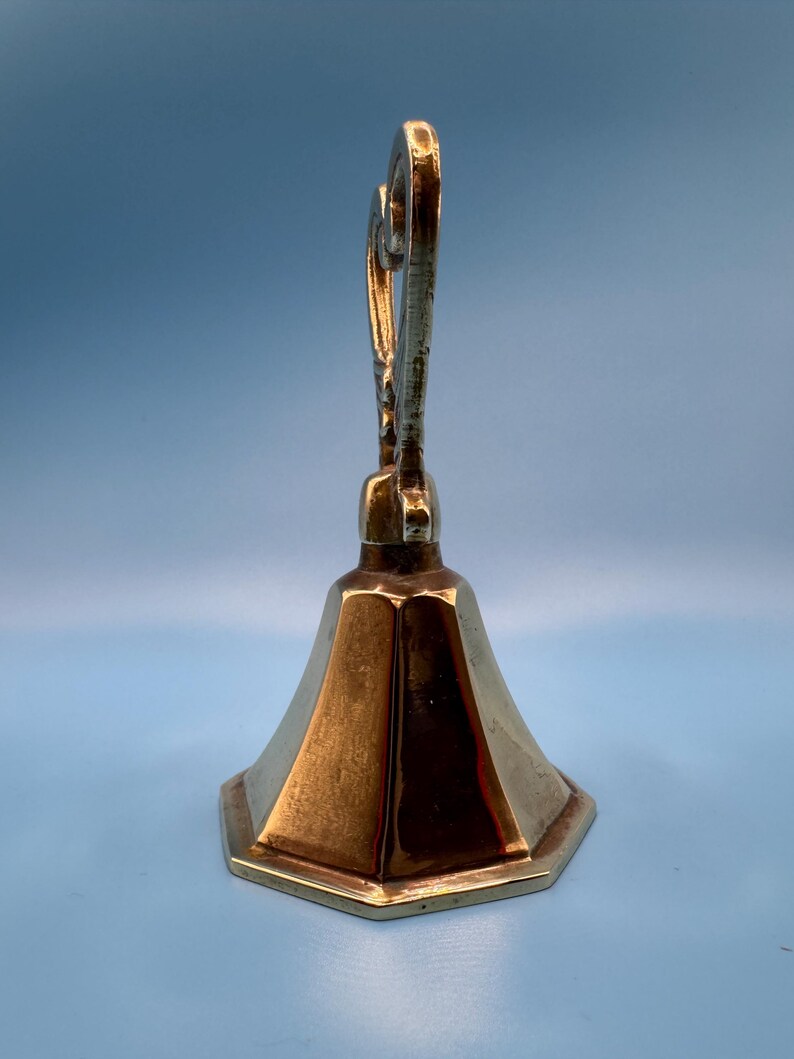K&ouml;nnte beinhalten: Ein Messing-Kerzenhalter mit sechseckigem Sockel und einem ausgestellten, facettierten K&ouml;rper. Die Oberseite weist ein verziertes, gerolltes Design auf. Der Kerzenhalter hat eine warme, goldene Farbe und steht vor einem hellblauen Hintergrund.