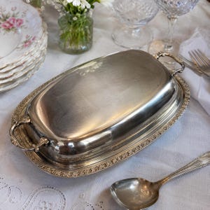 Op de afbeelding: Een zilverkleurige, ovale serveerschaal met deksel en sierlijke handgrepen. De schaal heeft een decoratieve rand en staat op een wit tafelkleed, samen met een lepel, borden en glaswerk. De scène suggereert een formele eetsituatie.