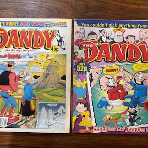 May include: Two vintage "Dandy" comic books. The left cover features "Desperate Dan" and the right cover has a variety of cartoon characters. The comics are brightly colored with text and illustrations, showcasing classic British humor.