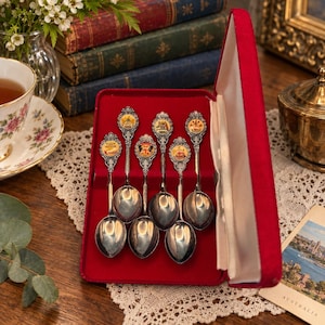 May include: A set of six silver-toned metal souvenir spoons arranged in a red velvet-lined presentation case. Each spoon features an ornate handle topped with a circular emblem displaying colourful crests and text representing various locations.