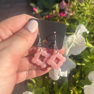 Könnte beinhalten: Ein Paar rosa, blütenförmige Ohrringe mit silberfarbenen Haken. Die Ohrringe werden vor einem schwarzen Etikett mit einem Hintergrund aus weißen Blumen und grünem Laub präsentiert.