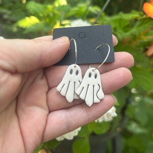 Peut inclure: Une paire de boucles d'oreilles en forme de fantôme blanc avec des détails noirs dessinés à la main pour les yeux et les plis du corps. Les breloques fantômes sont suspendues à de fins anneaux métalliques argentés fixés sur un support noir.