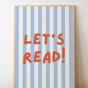 May include: A framed print with a light wood frame. The print features a blue and white striped background with the words "LET'S READ!" in orange letters. The artwork is displayed on a wooden floor.