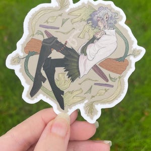 May include: A sticker featuring an illustration of a character with pale skin, gray hair, and pointed ears, wearing a white robe and black boots. The character is surrounded by green and brown plant-like elements. The sticker is held by a hand against a green background.