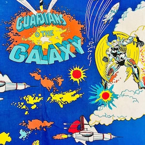 May include: Blue fabric with a comic book design. The words "GUARDIANS OF THE GALAXY" are in bold letters. The design includes rockets, explosions, and superhero figures in action, with a retro aesthetic.