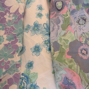 May include: Three distinct fabric patterns are displayed side by side. The left features purple and teal flowers on white. The centre shows blue flowers on white. The right displays large, muted floral shapes in shades of pink, blue, and green.