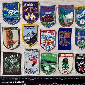 May include: A collection of fourteen embroidered fabric patches featuring New Zealand locations and symbols, including Fox Glacier, Auckland, Wellington, and Mount Cook, arranged next to a black metal ruler measuring up to 300 millimetres.