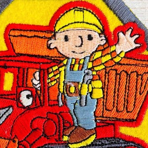 May include: Embroidered patch depicting a cartoon construction worker in a yellow hard hat and plaid shirt, waving. The patch includes a red truck and is set against a yellow background with a grey border. The design is detailed with various colours.