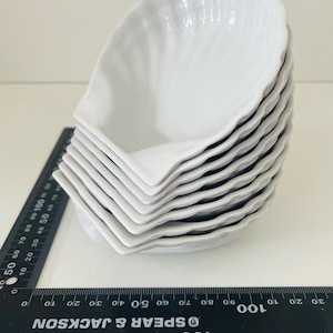 May include: A stack of eight white, leaf-shaped ceramic dishes. Each dish has a scalloped edge and a textured interior. A black ruler with white markings is in the foreground. The dishes are suitable for serving appetizers or small portions.