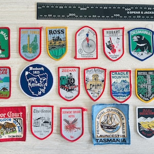 May include: A selection of embroidered patches depicting Tasmanian landmarks, including Cradle Mountain and Russell Falls. The patches are in various shapes and colors, with text identifying each location. A ruler, marked in centimeters, is placed next to the patches.
