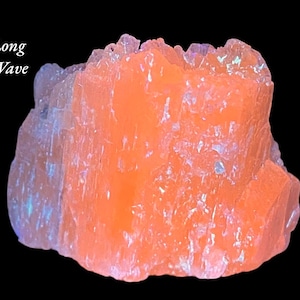 May include: A vibrant orange crystal formation against a black background. The crystal has a rough, textured surface with hints of blue and purple iridescence. The words "Long Wave" are in the upper left corner.