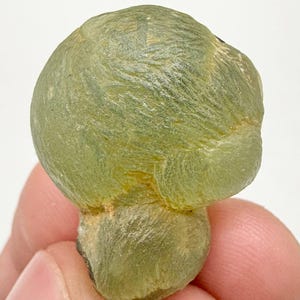 Puede incluir: Un espécimen mineral de color verde claro, de forma redondeada y bulbosa, con una superficie texturizada. La piedra tiene una base redondeada más pequeña. El color es verde translúcido con algunas zonas más oscuras.