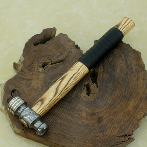 Könnte beinhalten: Ein handgefertigter Hammer mit Holzgriff und schwarzem Ledergriff. Der Hammerkopf hat ein gemustertes Metalldesign. Der Hammer wird auf einer dunklen Holzoberfläche präsentiert.