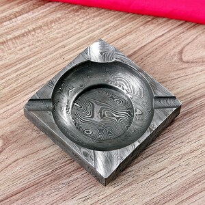 May include: Square ashtray with a distinctive, patterned design in shades of grey and silver. The ashtray has a circular indentation in the centre and grooves on the sides for cigarettes. The overall design is geometric and modern.