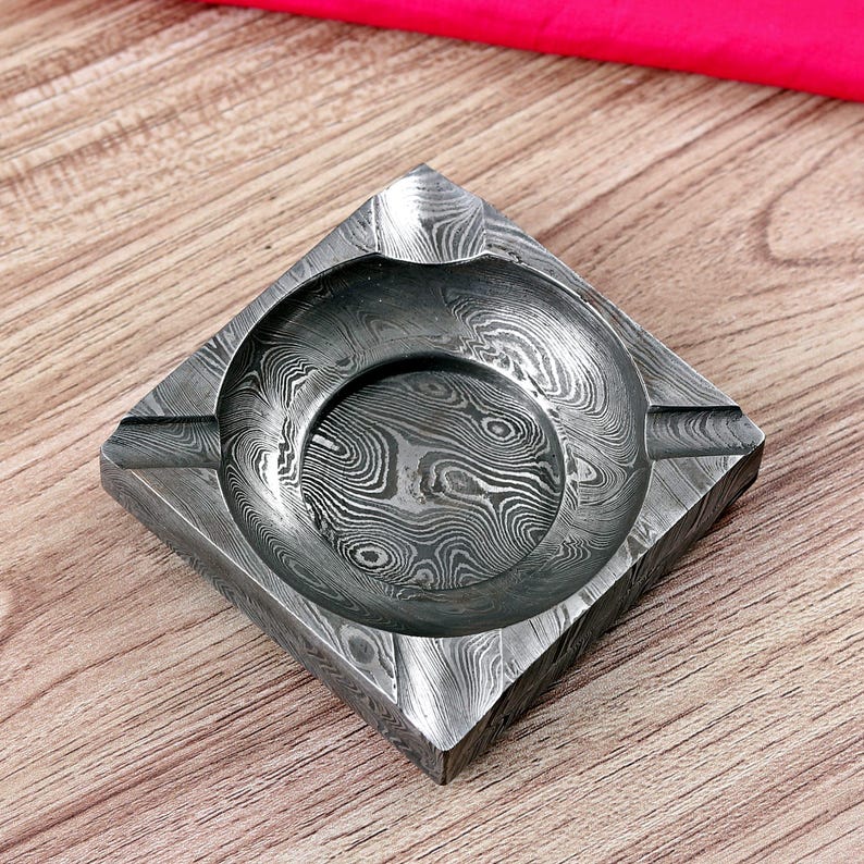 May include: Square ashtray with a distinctive, patterned design in shades of grey and silver. The ashtray has a circular indentation in the centre and grooves on the sides for cigarettes. The overall design is geometric and modern.