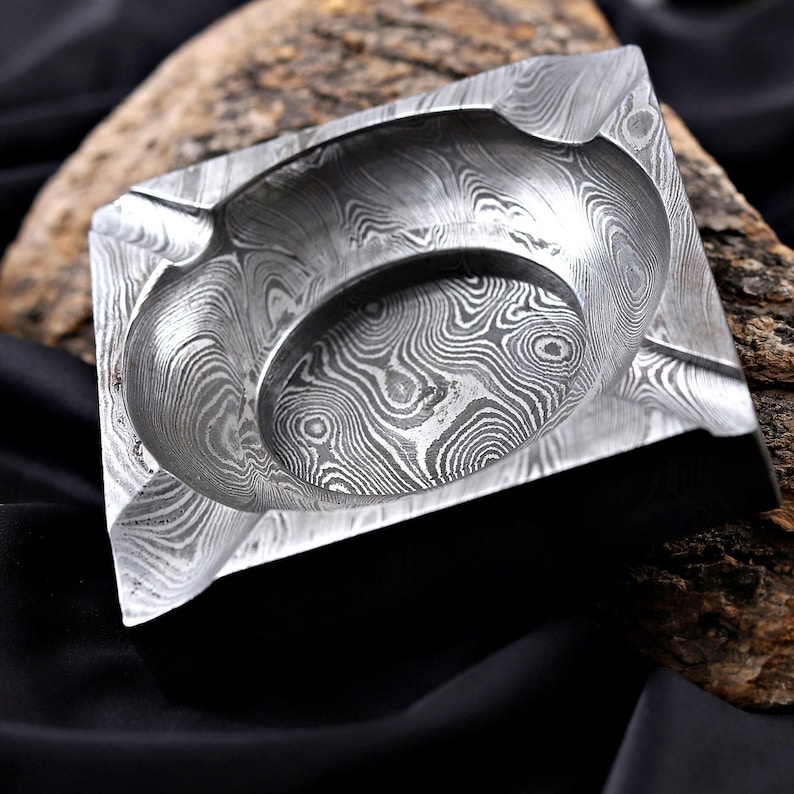 May include: Square metal ashtray with a distinctive, swirling pattern. The ashtray features a deep, circular indentation in the centre for holding cigars or cigarettes, and a surrounding lip for resting them. The metal has a silver colour.