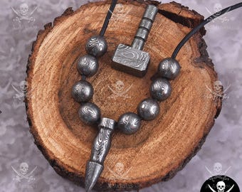 Puede incluir: Un conjunto de joyas de metal hechas a mano, que incluye un collar con cuentas estampadas y un colgante, y un martillo en miniatura. Las joyas se exhiben sobre una superficie de madera, mostrando los intrincados diseños y texturas del trabajo en metal.
