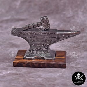 Może przedstawiać: Miniaturowy zestaw kowadła i młotka. Kowadło wykonane jest z metalu o wzorze i stoi na drewnianej podstawie. Młotek spoczywa na kowadle. Zestaw został zaprojektowany z dbałością o szczegóły i fakturę.