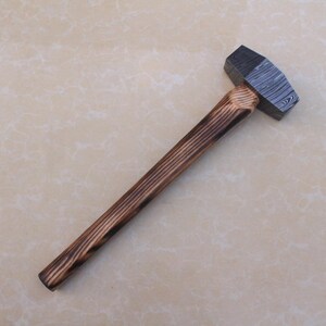 May include: A hammer featuring a patterned metal head and a long wooden handle. The handle has a dark, burnt wood grain pattern. The hammer is designed for striking and shaping materials.