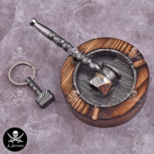 May include: A wooden ashtray with a metal insert and a matching hammer-shaped ashtray tool. A small hammer-shaped keyring is also included. The items have a patterned design and are set on a grey surface.