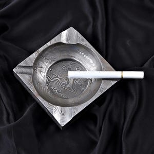 May include: Square metal ashtray with a distinctive patterned design. The ashtray features a circular indentation and a groove for a cigarette. A lit cigarette rests in the groove. The ashtray is on a black surface.
