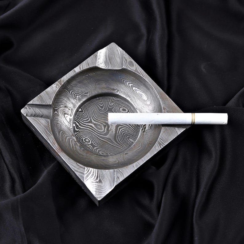 May include: Square metal ashtray with a distinctive patterned design. The ashtray features a circular indentation and a groove for a cigarette. A lit cigarette rests in the groove. The ashtray is on a black surface.