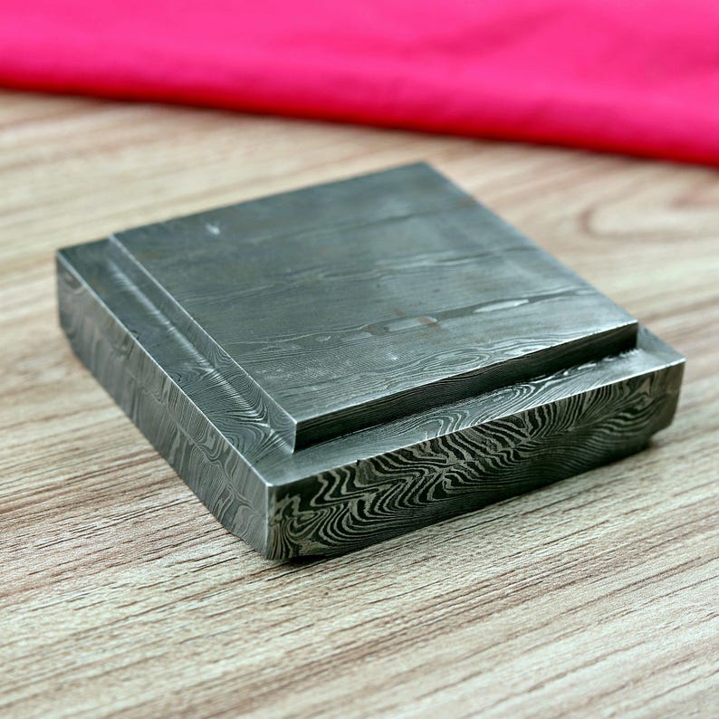 May include: A square, layered metal block with a distinctive wood grain pattern. The block is a dark grey colour and rests on a wooden surface. A pink fabric is in the background.