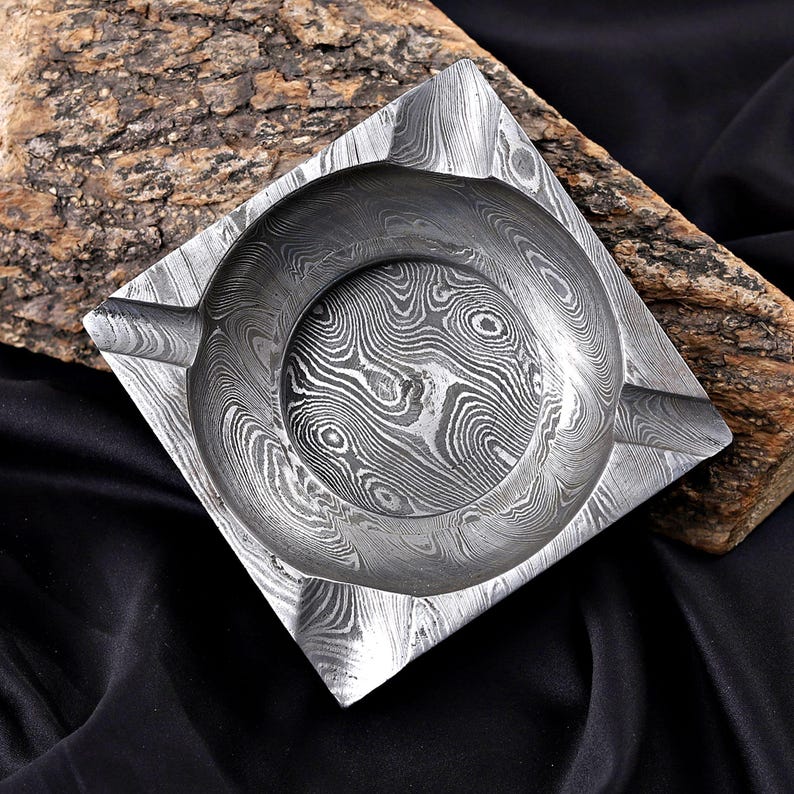 May include: Square metal ashtray with a circular centre and a textured, swirling pattern. The ashtray has a small rest for a cigarette. The ashtray is made of metal and is sitting on a piece of wood.