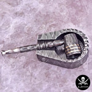 May include: A handcrafted, Damascus steel hammer with a textured handle and head, set within a matching U-shaped base. The hammer head features gold-colored stripes. The design showcases intricate patterns and a polished finish, ideal for blacksmithing or decorative purposes.
