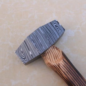 Può includere: Primo piano di un martello con una testa in metallo scuro a motivi e un manico in legno. La testa presenta un design a vortice simile alle venature del legno in tonalità di grigio e bianco. Il manico è in legno marrone chiaro con linee di venature marrone scuro.