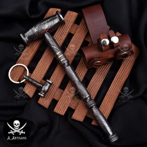 Puede incluir: Un conjunto de artículos de acero de Damasco, que incluye un bolígrafo, un llavero con forma de martillo y un abridor de botellas, expuestos sobre un soporte de madera. El bolígrafo presenta detalles dorados y una funda de cuero. Los artículos tienen un diseño distintivo.