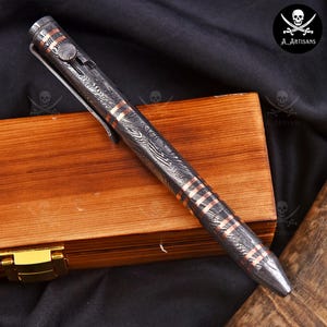 Op de afbeelding: Een Damascus stalen pen met koperen accenten ligt op een houten kist. De pen heeft een patroon met koperen banden. De pen heeft een clip en een drukknopmechanisme. Het is een schrijfinstrument.