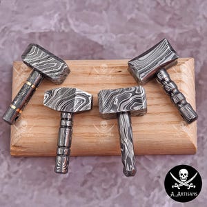 May include: Four handcrafted hammers with patterned metal heads and handles rest on a wooden surface. The hammers have a unique, swirling design in shades of gray and black. The handles vary in style, some with gold accents.
