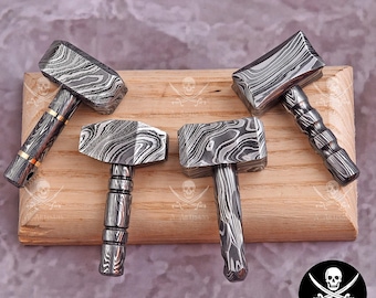 Puede incluir: Cuatro martillos artesanales con cabezas y mangos de metal estampados sobre una superficie de madera. Los martillos presentan un diseño arremolinado único en tonos grises y negros. Los mangos varían en estilo.
