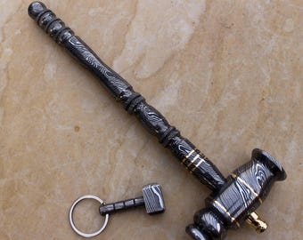 Puede incluir: Imagen detallada de un mazo y un llavero de mazo en miniatura. El mazo es negro con detalles dorados y un diseño estampado. El llavero es plateado con un pequeño dije de mazo. Ambos artículos están sobre una superficie marrón claro.