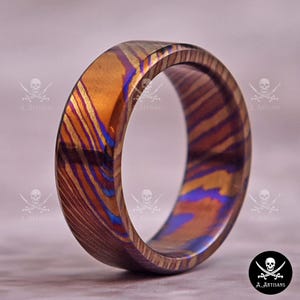 May include: A close-up of a colorful ring with a unique pattern. The ring features a blend of gold, purple, and blue hues, creating a striking visual effect. The ring's design showcases a textured, layered appearance, adding depth and character to the jewelry.
