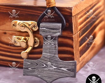 Puede incluir: Un colgante plateado con forma de martillo de Thor, con un patrón de vetas de madera. El colgante está suspendido de un cordón de cuero negro con un acento tejido de color marrón claro. El colgante está frente a una caja de madera con herrajes dorados.