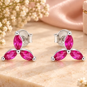 May include: A pair of silver stud earrings, each featuring three marquise-cut vibrant pink stones arranged in a floral-like cluster. The earrings have a polished metallic finish and secure post backs, showcasing a symmetrical design with pointed petals.