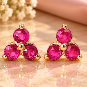 May include: A pair of stud earrings featuring three round, vibrant pink stones arranged in a triangular cluster set in gold-toned metal. Each earring showcases a trio of faceted gems that catch the light, creating a bright and reflective appearance.