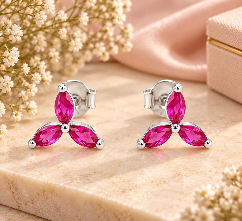 May include: A pair of silver stud earrings, each featuring three marquise-cut vibrant pink stones arranged in a floral-like cluster. The earrings have a polished metallic finish and secure post backs, showcasing a symmetrical design with pointed petals.