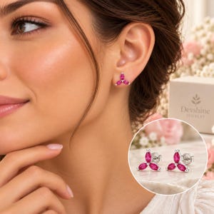 May include: A pair of silver stud earrings featuring three marquise-cut vibrant pink stones arranged in a floral cluster design. The earrings are shown worn on an earlobe and also displayed separately in a circular inset next to a Devshine Jewelry box.