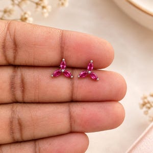 May include: A pair of stud earrings featuring three marquise-cut fuchsia pink stones arranged in a floral cluster pattern, set in a polished silver-toned metal base. Each earring consists of three petals meeting at a central point.