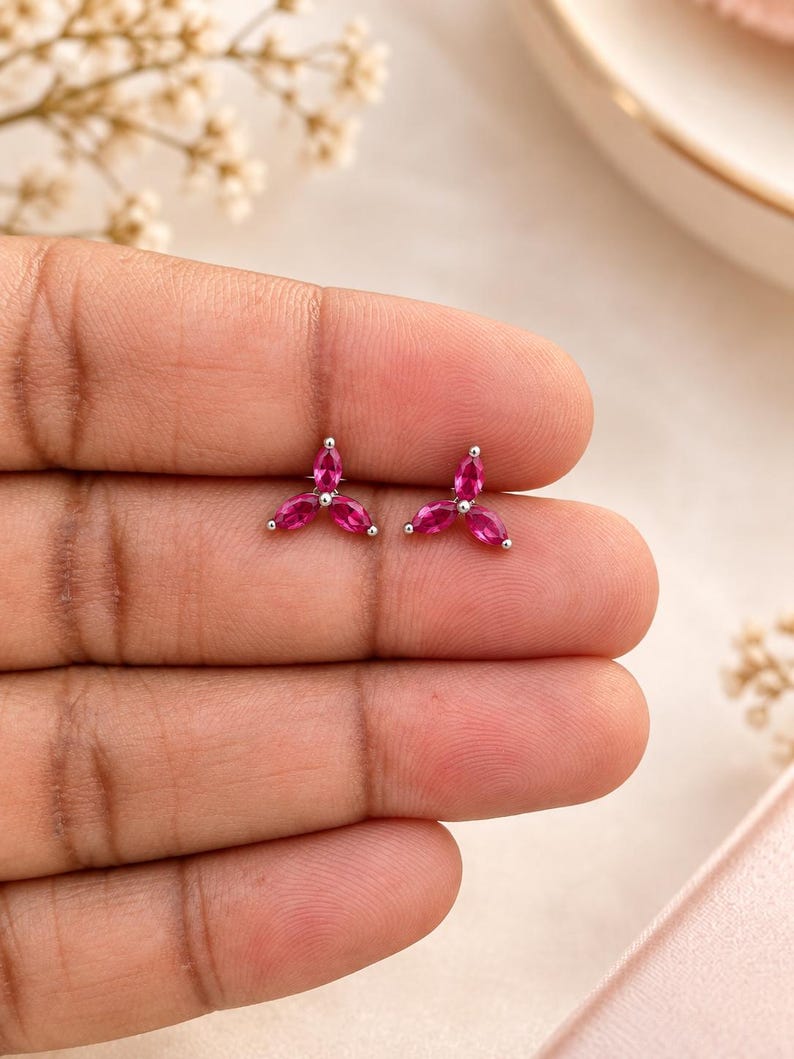 May include: A pair of stud earrings featuring three marquise-cut fuchsia pink stones arranged in a floral cluster pattern, set in a polished silver-toned metal base. Each earring consists of three petals meeting at a central point.