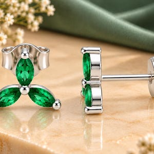 May include: A pair of silver stud earrings featuring three marquise-cut green stones arranged in a clover-like cluster. Each earring has a post back and a secure butterfly clutch, showcasing a polished metallic finish and vibrant green gemstones.