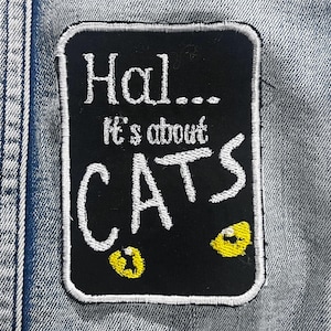 May include: A black rectangular patch with white embroidered text saying "Hal... It's about CATS." The patch has a white border and two yellow cat eyes. The patch is on a denim background.
